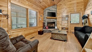 A cozy cabin interior with wooden walls, a stone fireplace, a flat-screen TV, and a comfortable seating area.
