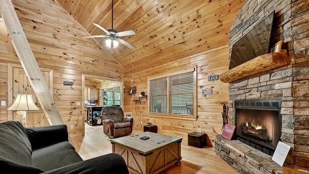 A cozy cabin interior with a stone fireplace, wooden walls, a ceiling fan, and a comfortable seating area.