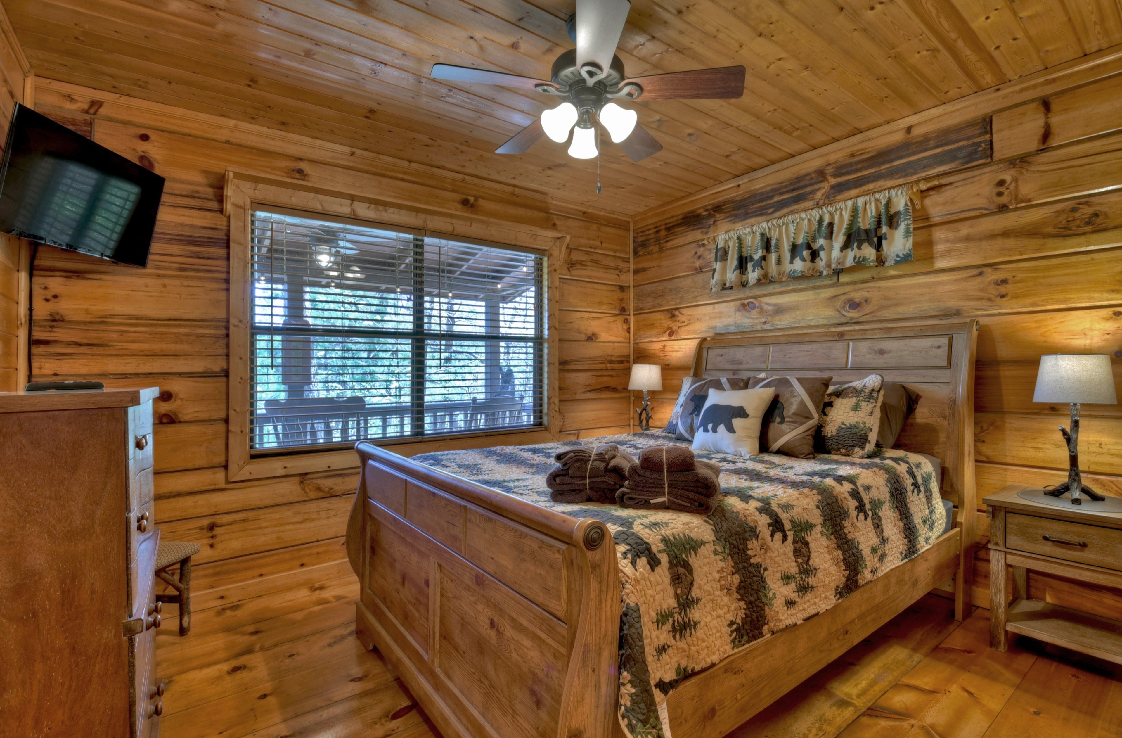 Une chambre dans une cabane en bois, avec un lit, un ventilateur de plafond, un téléviseur fixé au mur et une fenêtre avec des stores.