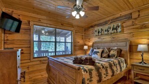 A wooden cabin bedroom with a bed, a ceiling fan, a TV mounted on the wall, and a window with blinds.