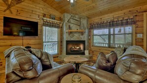 A cozy cabin interior with a stone fireplace, leather sofas, a television mounted on the wall, and wooden walls.