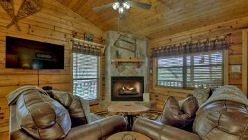 A cozy cabin interior with a stone fireplace, leather sofas, a television mounted on the wall, and wooden walls.