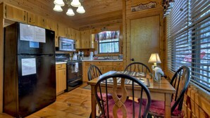 A wooden cabin interior with a kitchen featuring a refrigerator, microwave, stove, and wooden cabinets. There is a dining table with chairs and a window with blinds.