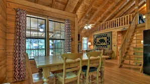 L’intérieur d’une cabane en bois comprend un coin repas, un escalier et une grande fenêtre avec des rideaux.