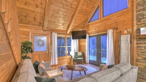 L’intérieur d’un chalet confortable, avec un grand plafond en bois, un canapé, un fauteuil et un téléviseur.