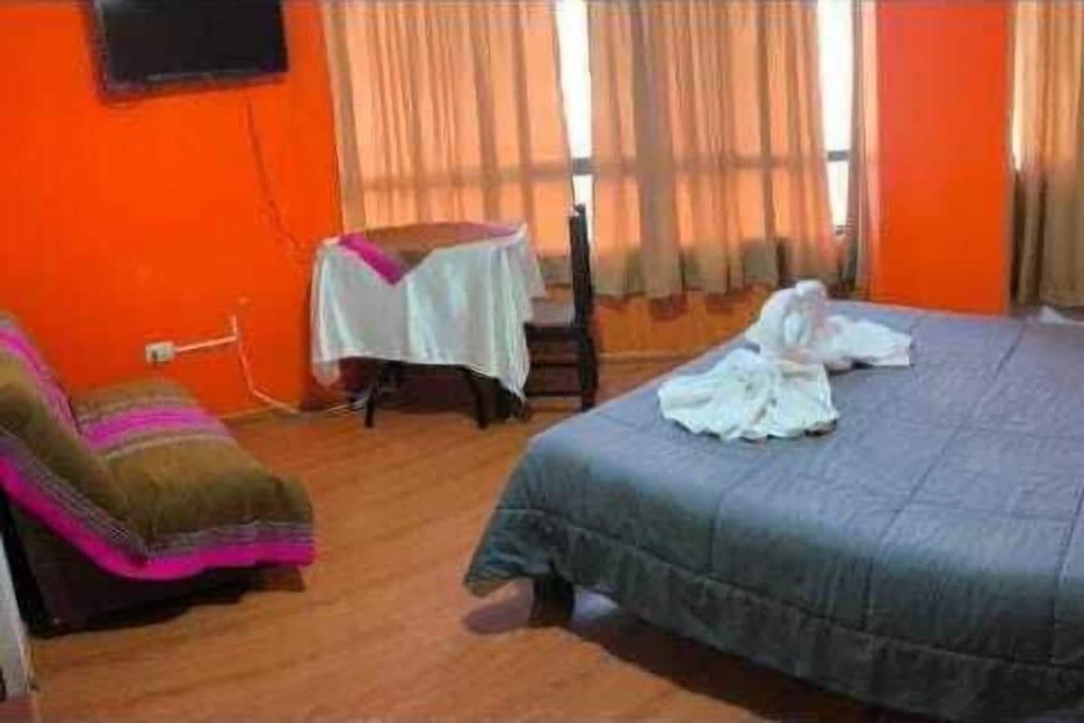 Quarto de hotel com cama, uma cadeira, uma mesinha e cortinas.