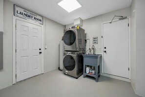 A self-service laundry room with a stacked washer and dryer, a sink, and a sign indicating the room is open 24 hours.