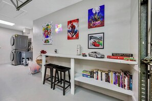 Une pièce avec un mur blanc, une longue table blanche, deux tabourets noirs, une bibliothèque remplie de divers ouvrages et un panneau lumineux au néon.