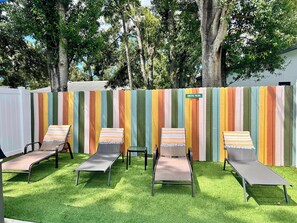 A row of four sun loungers with striped towels, a colorful striped fence, and a sign that reads "COASTAL PALMS" in a garden setting.