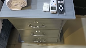 A small, light-colored wooden dresser with three drawers, a rolled white towel, a white box labeled "SENSY," and a remote control on top. - Hotel Pasarela Cali (Cali)