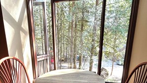 A round table with a wooden top and metal base, flanked by two wooden chairs with curved backs. The table is positioned in a room with large windows that offer a view of a forested area outside. - Mountain Retreat near mt Baker, cozy 1bedroom Condo w/ Pool, Hot Tub, Crib, wifi (Deming)