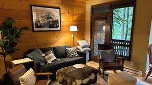 A cozy log cabin interior with a sofa, chairs, a table, and a large window offering a view of trees. - Mountain Retreat near mt Baker, cozy 1bedroom Condo w/ Pool, Hot Tub, Crib, wifi (Deming)
