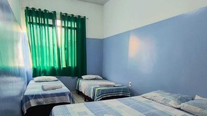 Quarto com três camas de solteiro, cada uma com colcha azul e branca estampada e travesseiros brancos. Há uma cortina verde na janela, e um ventilador de teto é visível.