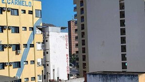 Um hotel com um letreiro chamativo 'HOTEL' em um edifício branco, rodeado por outros edifícios, incluindo um com a placa 'TE CARMELO'.