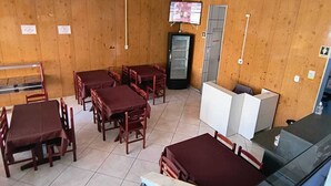 Quarto com paredes de madeira, chão de azulejos e várias mesas com cadeiras. Há uma televisão fixada na parede e um frigorífico.