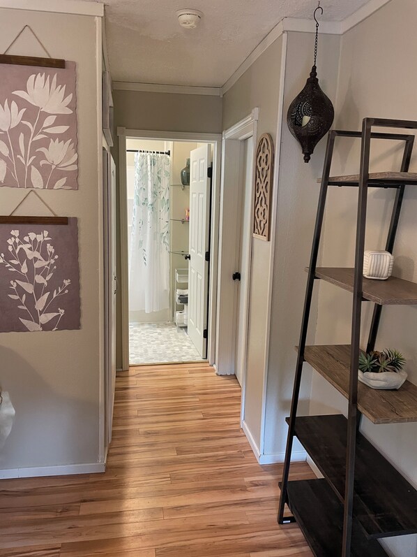 A hallway with wooden flooring, a decorative wall hanging, a metal shelf unit, and a door leading to a bathroom.