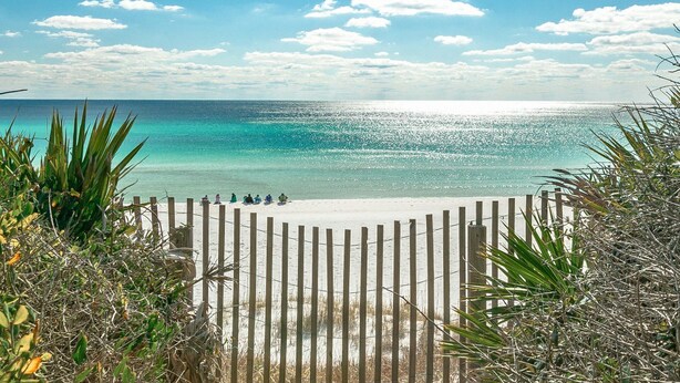 Ein Strand mit einer Holzlattenveranda, klarem türkisblauem Wasser und einigen Personen, die am Sand sitzen.