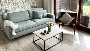 A modern living room with a grey sofa, a coffee table, and a view of a garden through a sliding glass door.