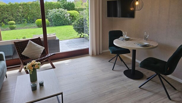 A modern living room with a large sliding glass door leading to a garden, a round dining table with chairs, a flat-screen TV mounted on the wall, and a coffee table with a vase and candles.