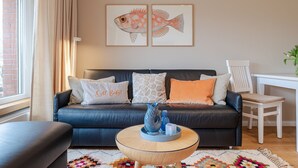 Ein Wohnzimmer mit einem dunkelbraunen Ledersofa, einem runden Holztisch und einem farbenfrohen Teppich. An der Wand befinden sich zwei Bilder von Fischen in Rahmen, ein Sessel und ein kleiner Tisch mit einer Pflanze.