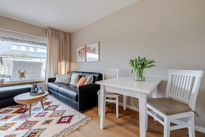 Sala de estar com sofá de couro preto, mesa de jantar branca e cadeira branca. Há um tapete colorido no chão, uma janela com cortinas e um vaso com plantas verdes na mesa.