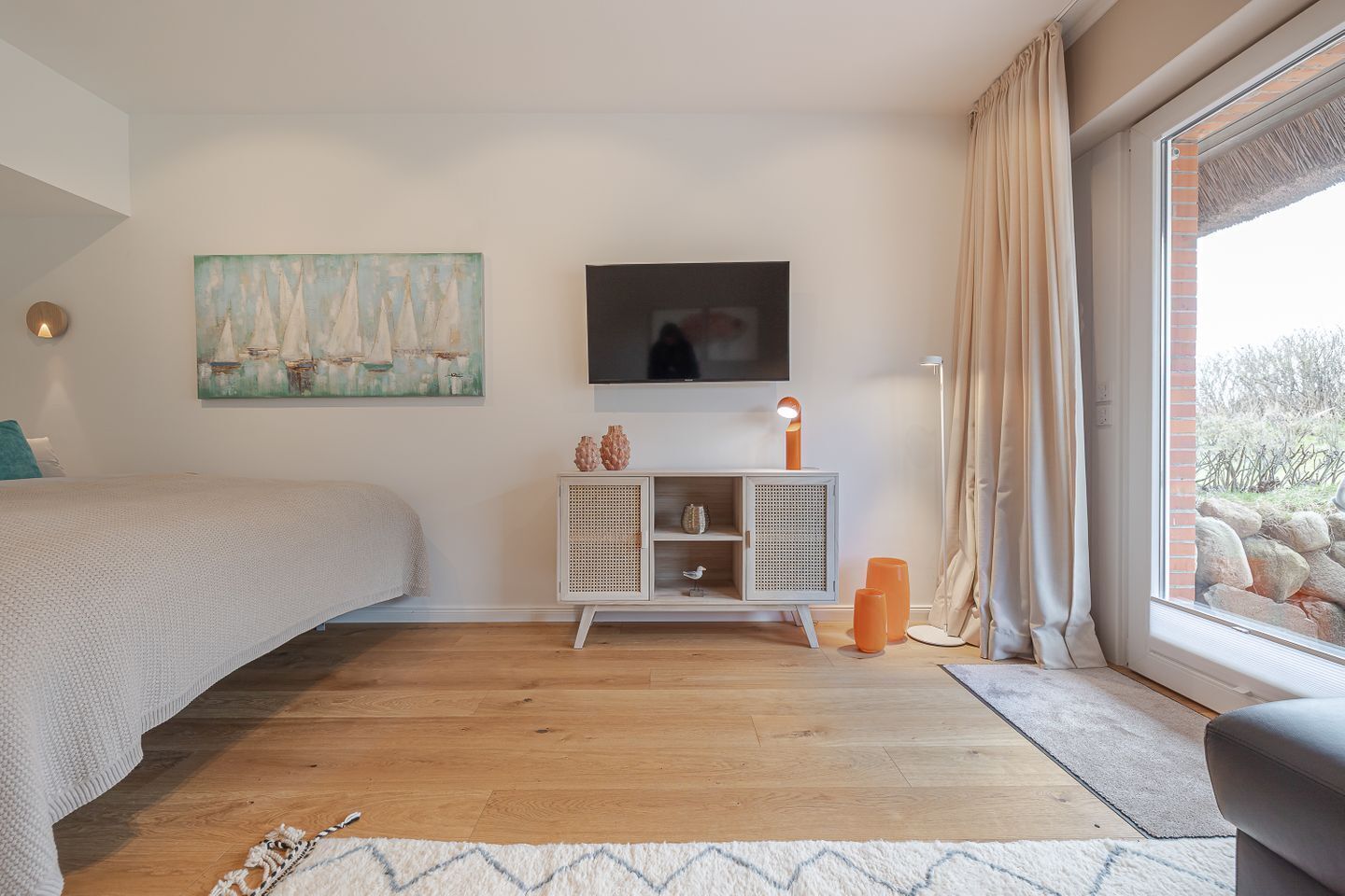 A bedroom with a bed, a TV mounted on the wall, a small cabinet, and a window with curtains.