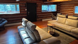 A log cabin interior with a sofa, a chair, and a small table. There are two windows providing views of the outside. - Hideout in the Mountains HOT TUB and River access (Gallatin Gateway)