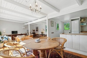 Ein geräumiges Zimmer mit einem runden Holztisch, Korbstühlen, einer Kronleuchter, weißen Schränken und Blick ins Grüne.