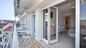Un balcon avec une table et des chaises, une porte-fenêtre coulissante donnant sur une chambre, et une vue sur le paysage urbain.