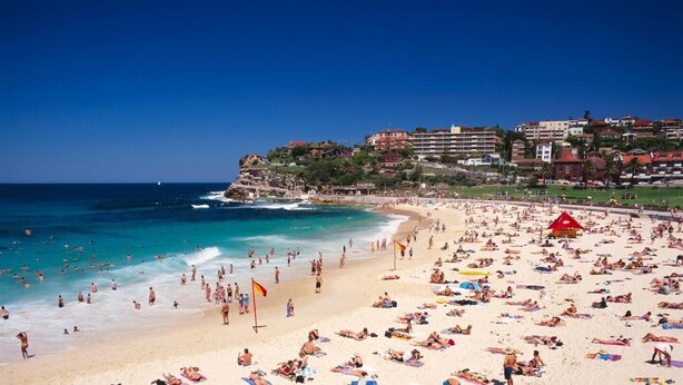 Una playa concurrida, gente tomando el sol y nadando, una bandera roja y edificios al fondo.