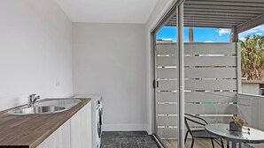 Un balcón con puerta de cristal corrediza, una pequeña mesa con sillas y una zona de cocina con lavabo y lavadora.