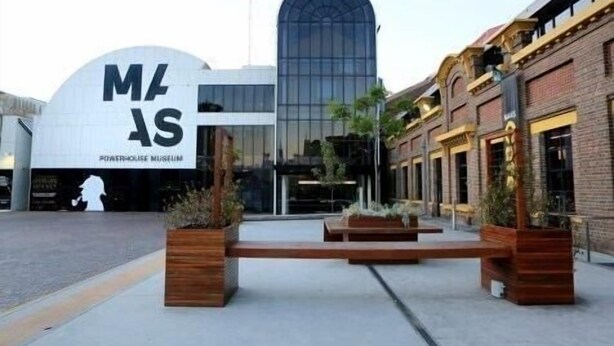 Un edificio moderno con un gran letrero con las letras 'M' y 'S', probablemente un museo, con fachada de vidrio y un edificio de ladrillo tradicional cercano.
