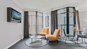 A modern living room with a flat-screen TV, a round coffee table, two armchairs, a small table with a vase, and a large window with curtains.