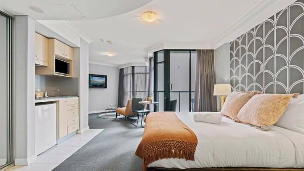 A modern hotel room with a bed, a desk, and a kitchenette.