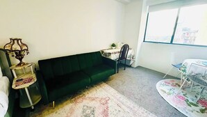 Ein Wohnzimmer mit einem grünen Sofa, einem kleinen Tisch mit einer Lampe, einem Sessel und einem Fenster mit Blick auf Gebäude.