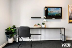 Moderni toimistotila, jossa on työpöytä, tuoli, ruukkukasveja, seinälle asennettu televisio ja kehystetty kuva seinällä.