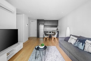 A modern living room with a flat-screen TV, a grey sofa, and a dining area with a table and chairs. - KozyGuru | Collingwood | Stylish 1 Bed Apt (Collingwood)