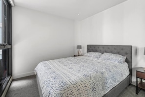 A bedroom with a large bed, a bedside table with a lamp, and a window with blinds. - KozyGuru | Collingwood | Stylish 1 Bed Apt (Collingwood)