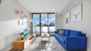 Una sala de estar moderna con un sofá azul, un mueble de televisión de madera y un amplio ventanal con vistas al cielo.