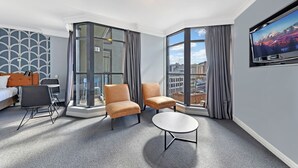 A hotel room with a bed, two armchairs, a small table, a TV, and large windows with curtains.