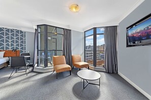 A hotel room with a bed, two armchairs, a small table, a TV, and large windows with curtains. - KozyGuru | Sydney CBD | Best Location Studio (Haymarket)