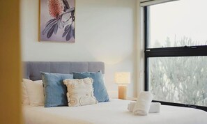 A bedroom with a bed, pillows, a nightstand, a lamp, and a window with a view.
