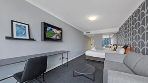 A modern hotel room with a bed, a desk, a chair, a TV, and a sofa. - KozyGuru | Sydney CBD | Prefect Location Studio (Haymarket)