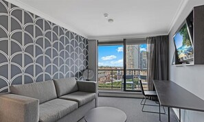 A modern hotel room with a grey patterned wall, a grey sofa, a small round table, a TV on the wall, and a balcony with a city view.