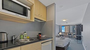 A modern kitchen with a microwave, sink, and kettle, a small dining area, and a bedroom with a bed and a view of the city. - KozyGuru | Sydney CBD | Prefect Location Studio (Haymarket)