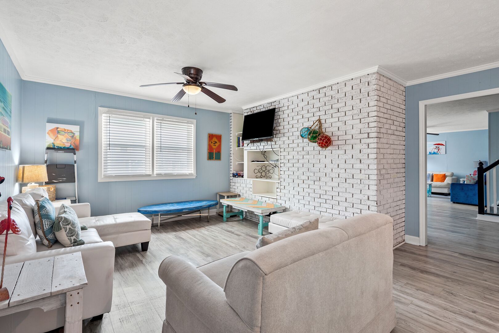 Ein Wohnzimmer mit blauer Wand, einer Akzentwand aus weißen Ziegelsteinen, einem Deckenventilator, einer blauen Bank, einem weißen Sofa, einem weißen Couchtisch und einem an der Ziegelwand montierten Fernseher.