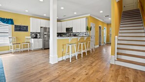 A spacious kitchen with white cabinets, a stainless steel refrigerator, and a microwave. There are three white bar stools and a chalkboard on the wall. - Luxury home w/private pool, walking distance to the beach, eateries and shopping (North Myrtle Beach)