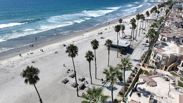 A beach with palm trees, a sandy shore, and a row of buildings with balconies. - Oceanfront 2BR/2BA Condo in Oceanside with Amazing Beach Access has AC! (Oceanside)