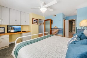 A bedroom with a bed, a desk with a television, a ceiling fan, and a door leading to a bathroom.