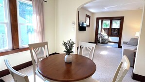 Un coin repas avec une table ronde en bois, quatre chaises blanches, un lustre et une vue sur le salon, accessible par une arche.